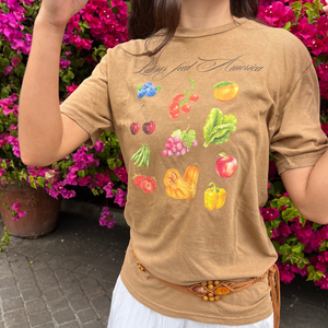 Person wearing a tan t-shirt that says Latinos Feed America with colorful fruit and vegetable graphics in front of pink flowers.