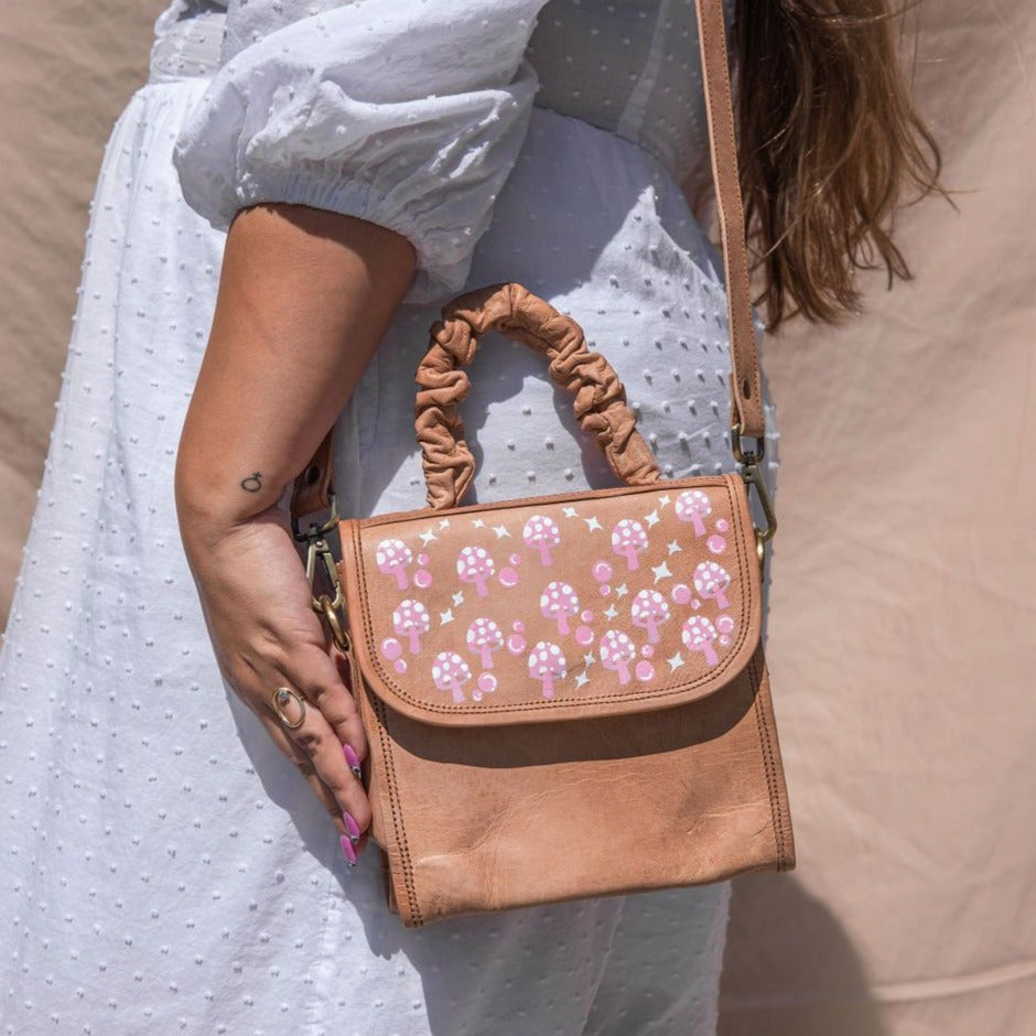 Brown leather purse with pink mushroom painting on front strap. With a scrunchie handle and a crossbody strap. Height is 7.5 inches,  Length is 7.5 inches, Width is 5 inches.