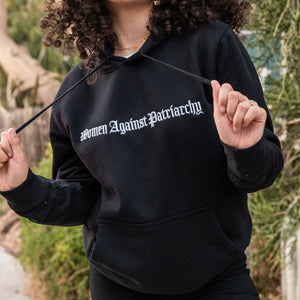 Model is wearing 'Women Against Patriarchy' black unisex fit sweatshirt. A heavy weight hoodie with kangaroo pocket. Sizes range from XS-3XL. Colors are Black or Tan.