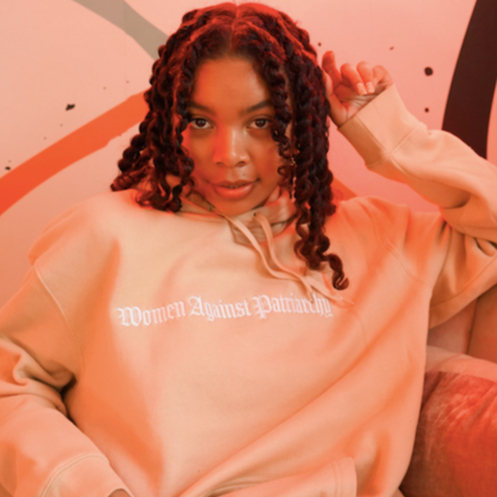 Model is wearing 'Women Against Patriarchy' tan unisex fit sweatshirt. A heavy weight hoodie with kangaroo pocket. Sizes range from XS-3XL. Colors are Black or Tan.